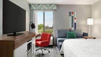 Habitación de hotel con una cama grande, un escritorio con televisor, una silla roja, un sofá gris y una ventana con vistas a zonas verdes y edificios.