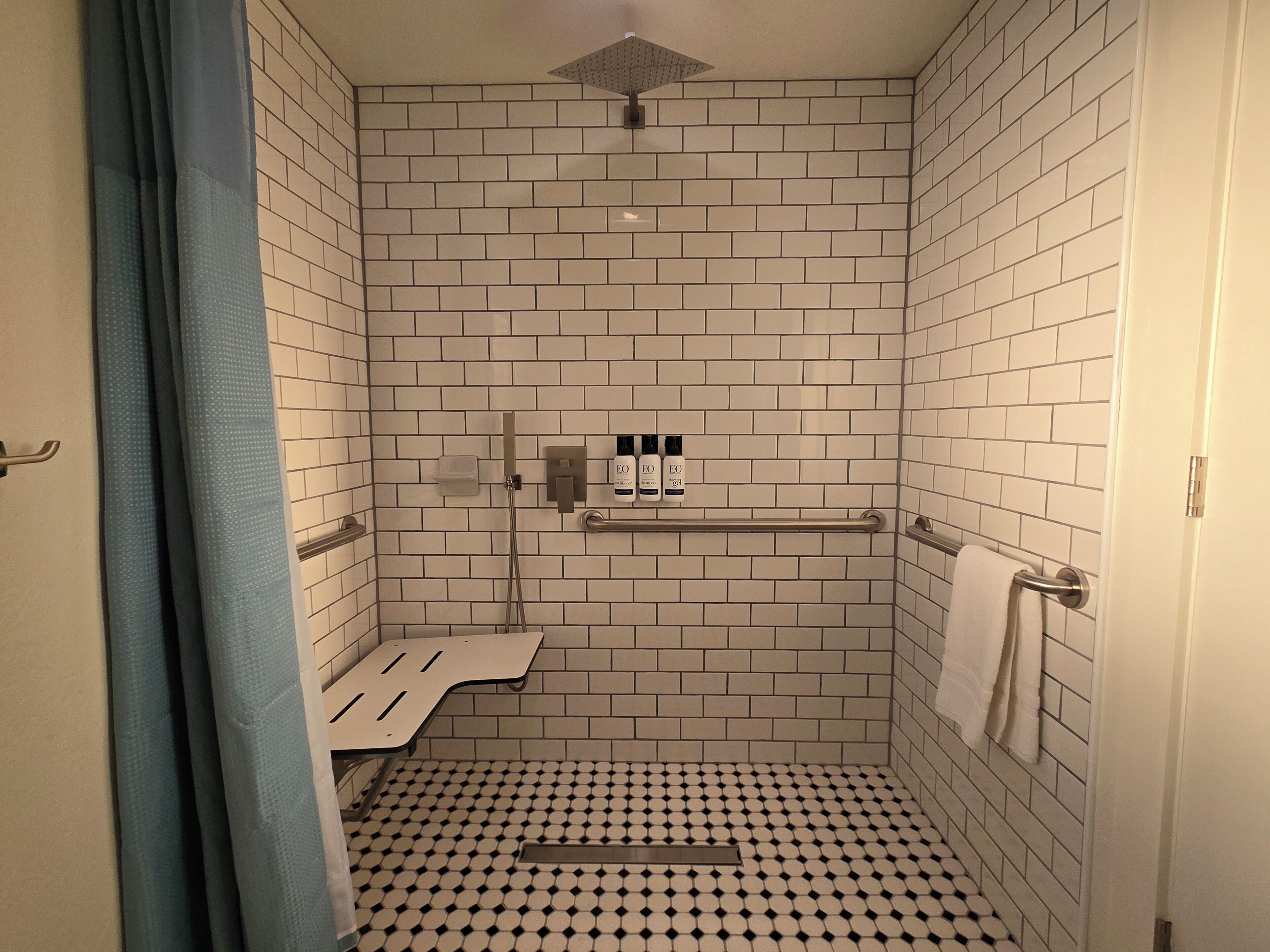 A small, tiled shower room with a showerhead, a towel rack, and a shelf with toiletries.