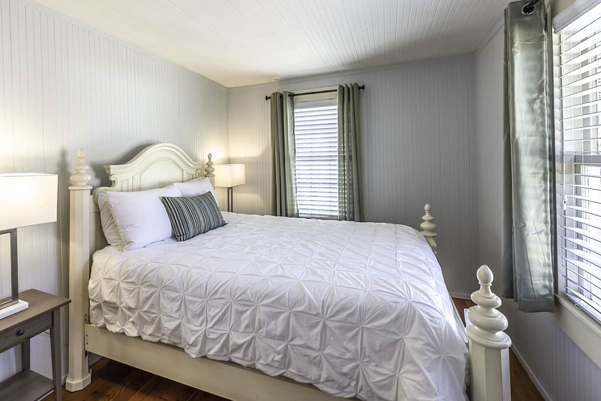 A neatly made bed with a white bedspread and pillows, a bedside table with a lamp, and a window with curtains.
