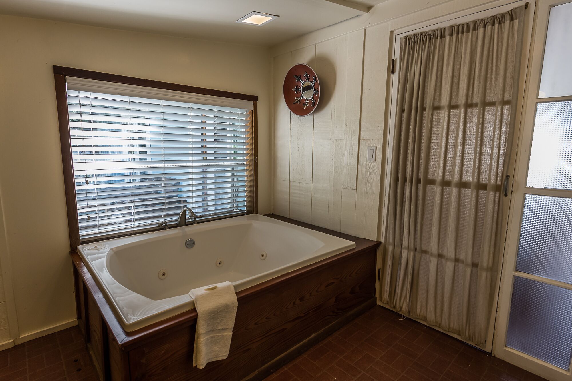 A bathroom with a large bathtub, a window with blinds, a door with a curtain, and a wall-mounted clock.
