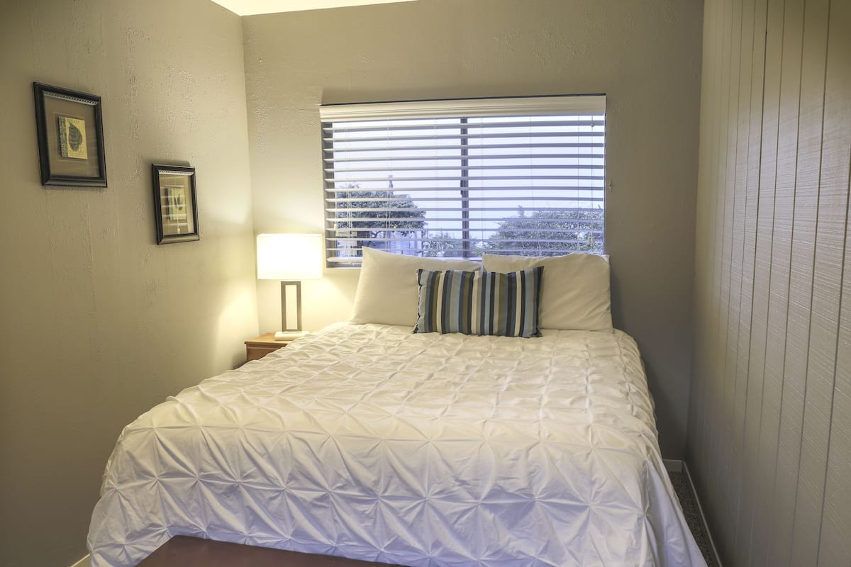 A bedroom with a large bed, a window with blinds, and two framed pictures on the wall.
