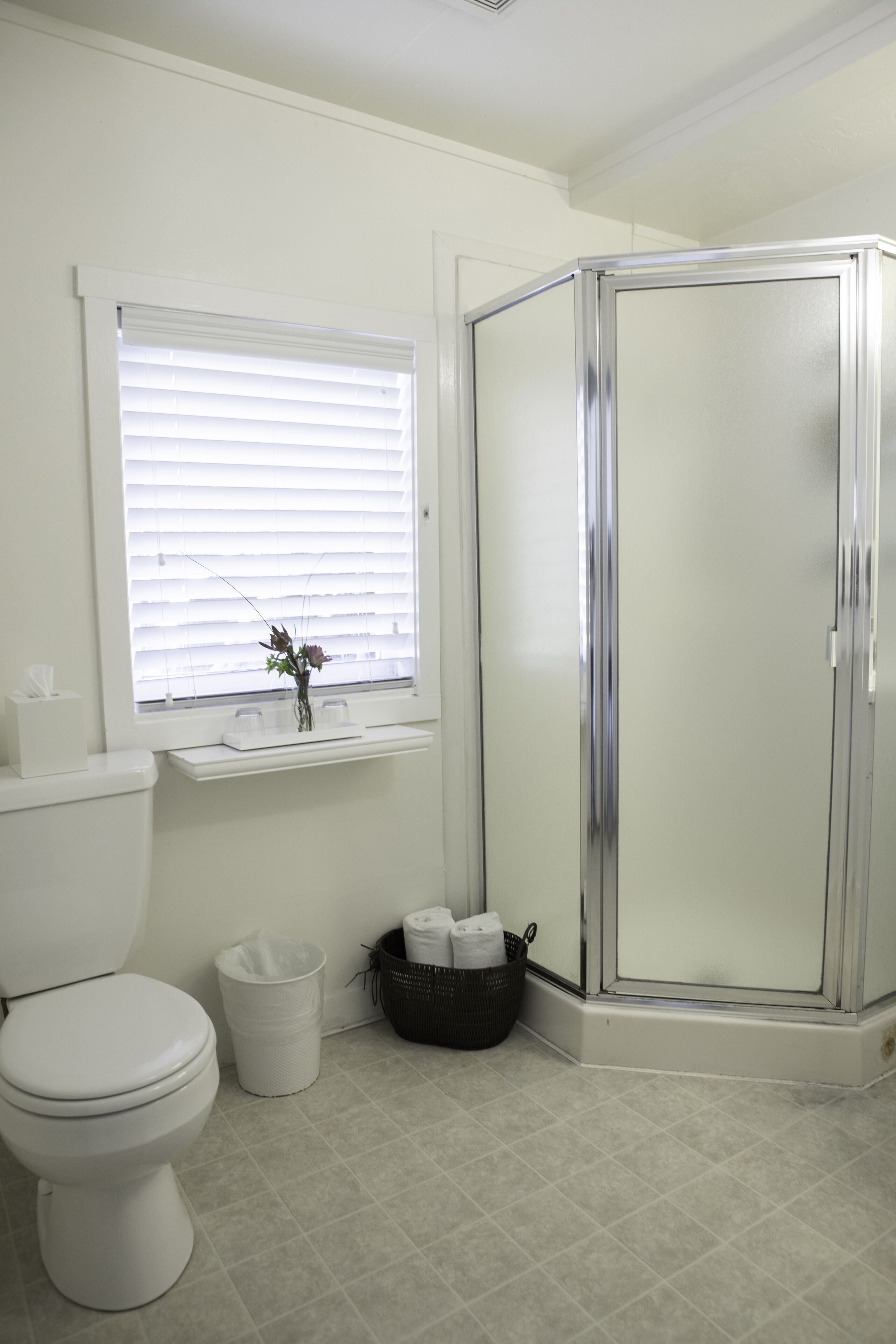 Un baño con inodoro, ventana con persianas, cabina de ducha y cubo de basura.