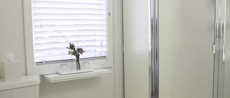 Un baño con inodoro, ventana con persianas, cabina de ducha y cubo de basura.