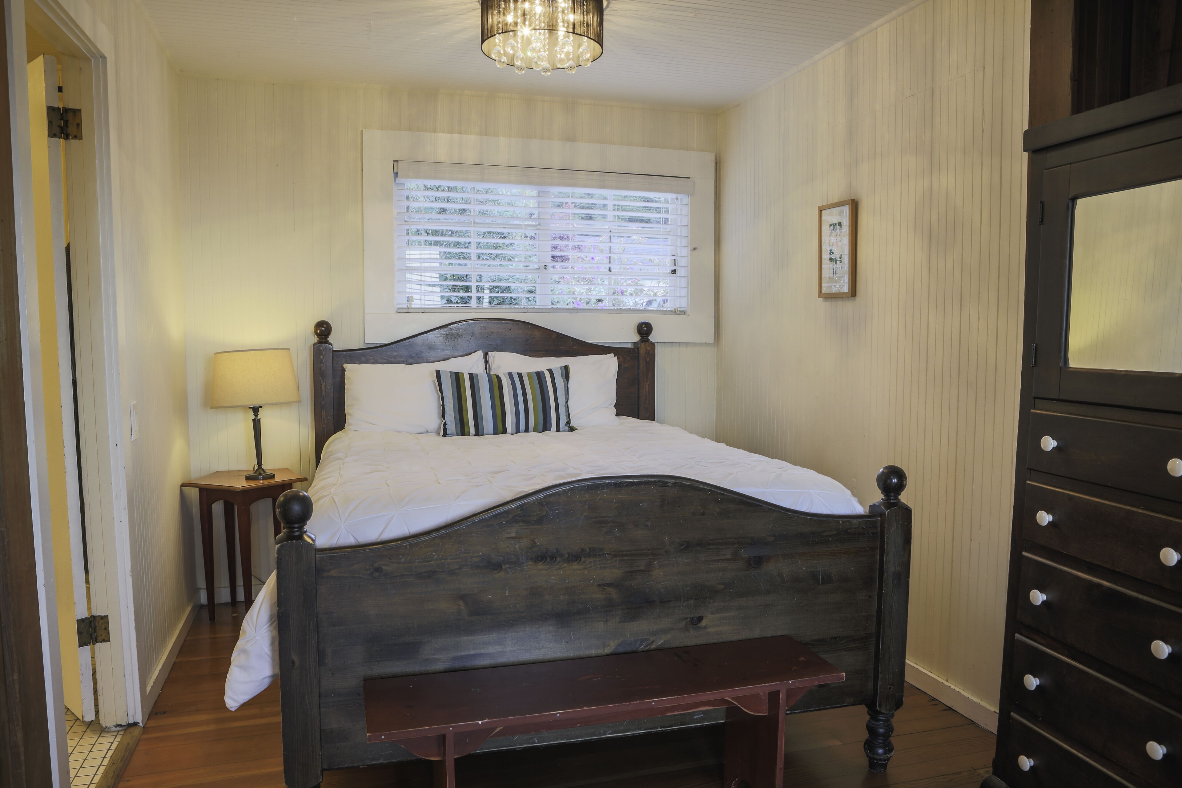 A bedroom with a wooden bed, a bedside table with a lamp, a window with blinds, and a dresser.