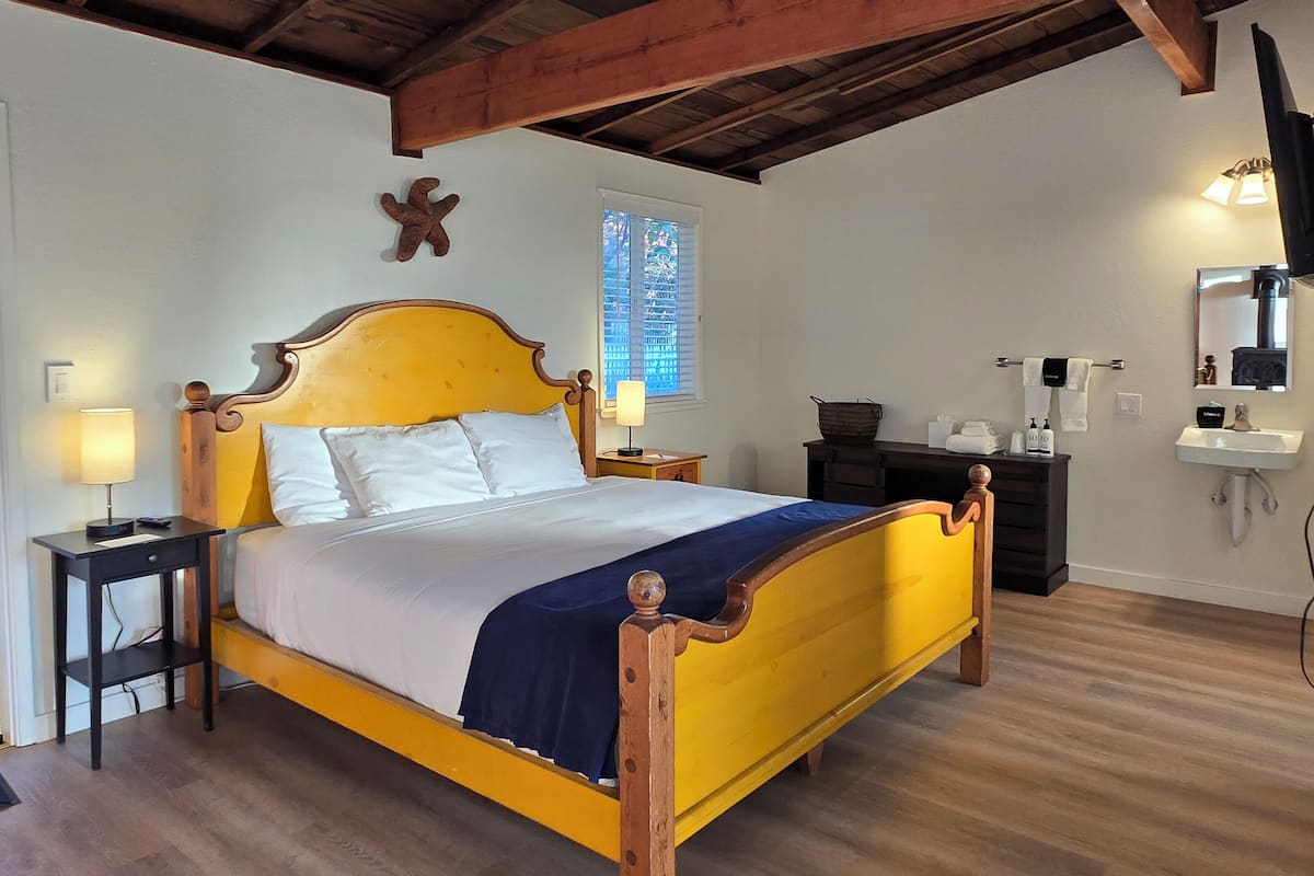 A bed with a wooden frame and a yellow headboard, a nightstand with a lamp, a window with blinds, a mirror, and a sink with toiletries.
