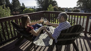 Dos personas sentadas en una mesa de madera en una terraza con barandilla, sosteniendo copas de vino.