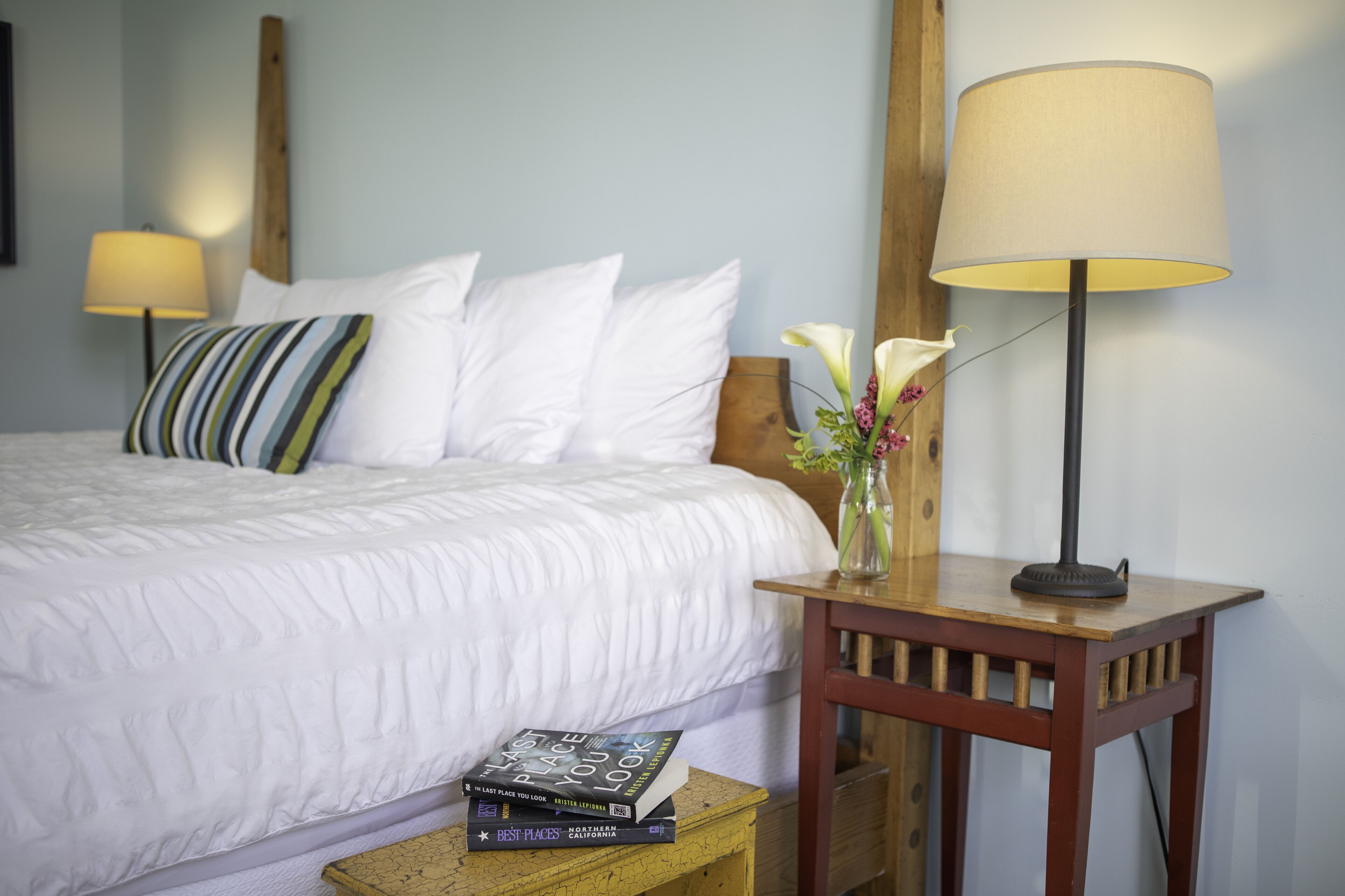 A neatly made bed with white bedding, a striped pillow, and a bedside table with a lamp and a vase with flowers.