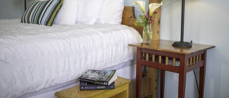 A neatly made bed with white bedding, a striped pillow, and a bedside table with a lamp and a vase with flowers.