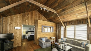 A cozy cabin interior with wooden walls, a television, a sofa, a dining table, and a window with blinds. - The Inn at Schoolhouse Creek (Little River)