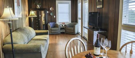 A cozy living room with a sofa, chairs, a dining table with a bottle of wine and glasses, and a TV.