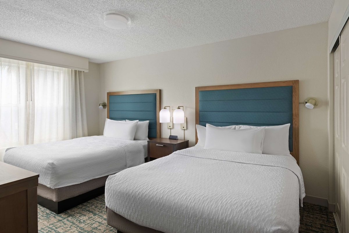 Habitación de hotel con dos camas, una mesita de noche, una lámpara y un ventanal con cortinas.
