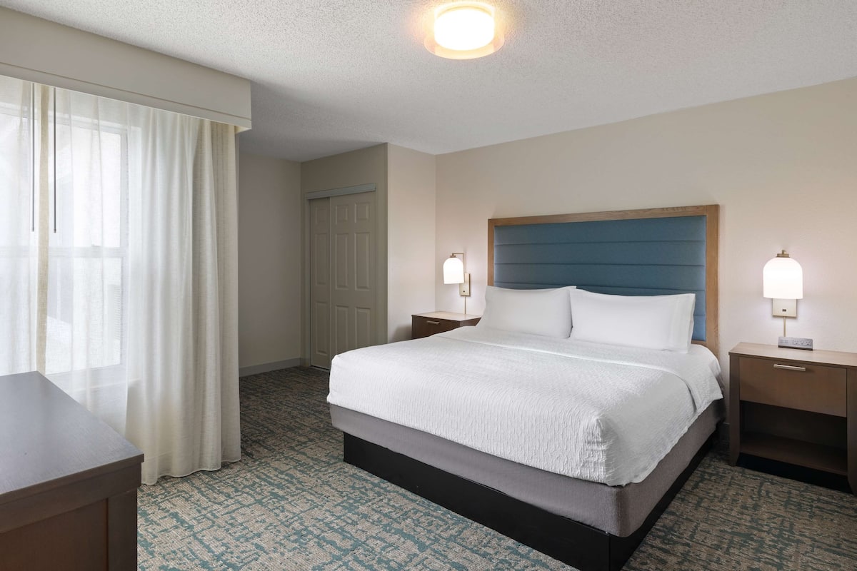 Habitación de hotel con una cama grande, dos mesitas de noche con lámparas, un ventanal con cortinas traslúcidas y una alfombra estampada.