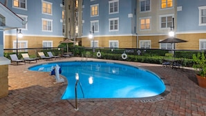 A swimming pool with lounge chairs and umbrellas, surrounded by a brick patio and a building with multiple windows.
