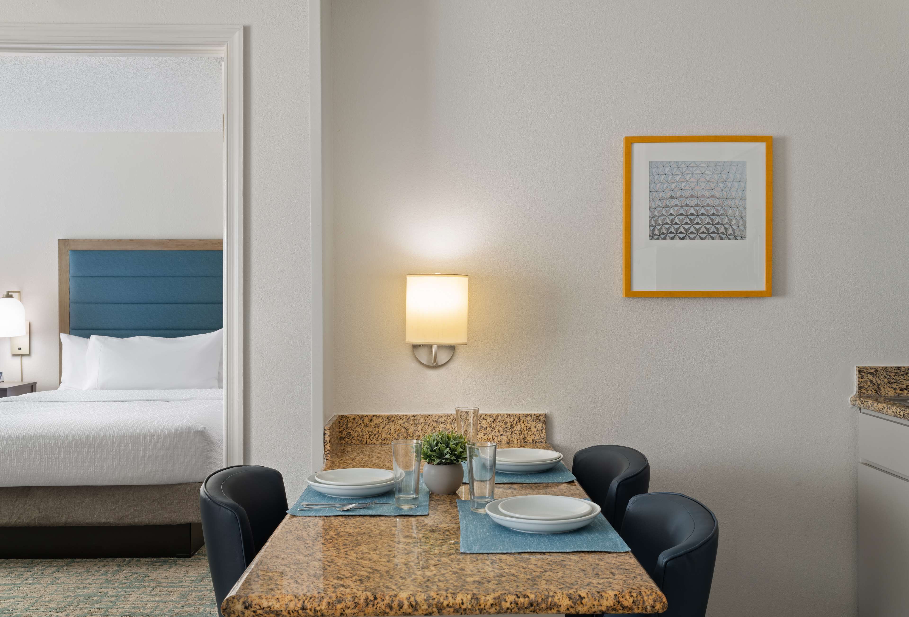 Habitación de hotel con cama, una mesa de comedor con platos y vasos, una lámpara de pared y un cuadro en la pared.