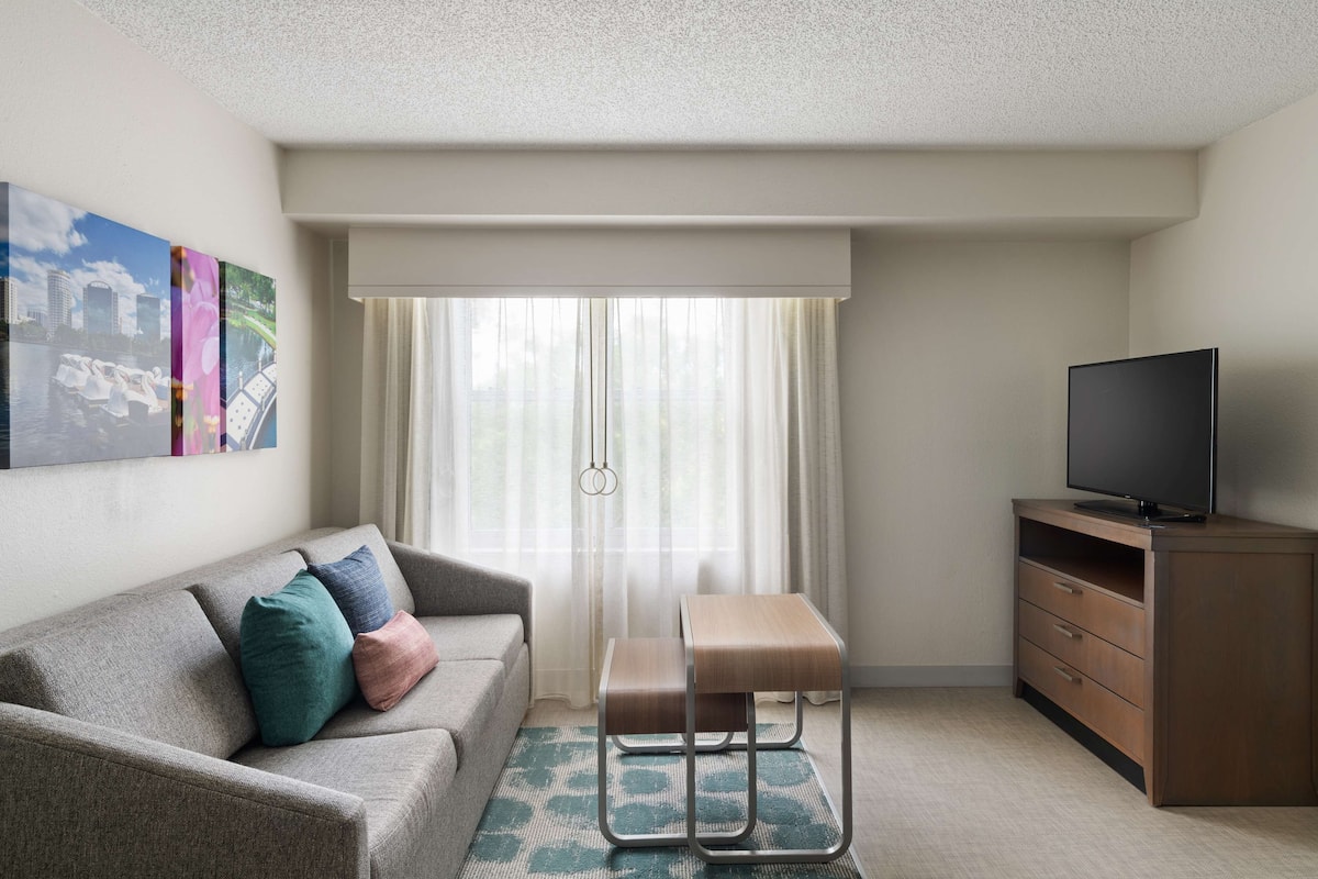 Una habitación de hotel moderna con sofá, una mesita de madera, televisión y ventana con cortinas traslúcidas.