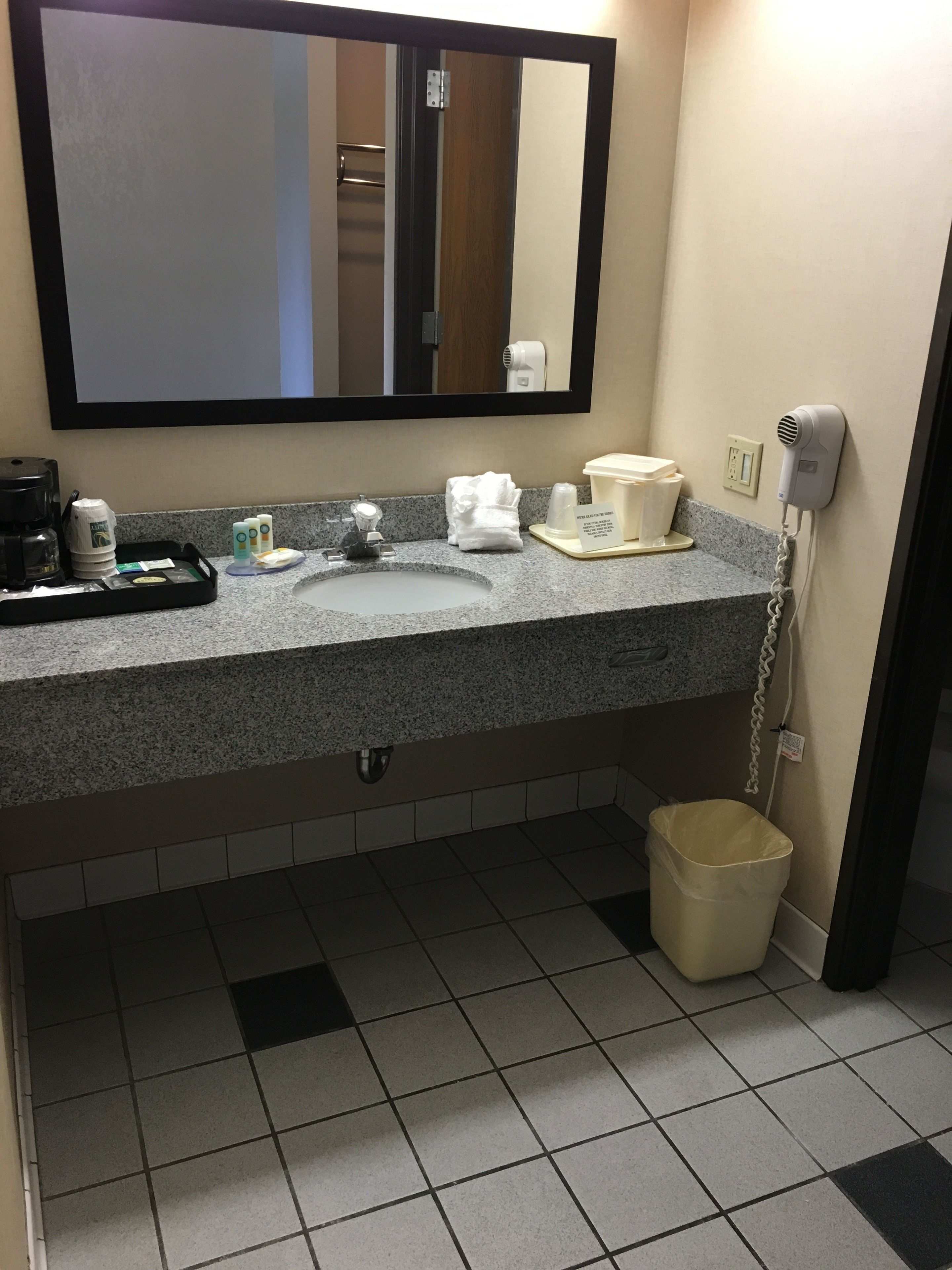 Casa de banho de hotel com bancada de granito, pia, espelho, secador de cabelo e lixeira.