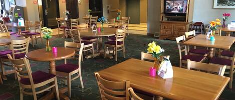 A hotel dining area with multiple wooden tables and chairs, some with purple cushions. There are vases with flowers on the tables, and a television is mounted on the wall.