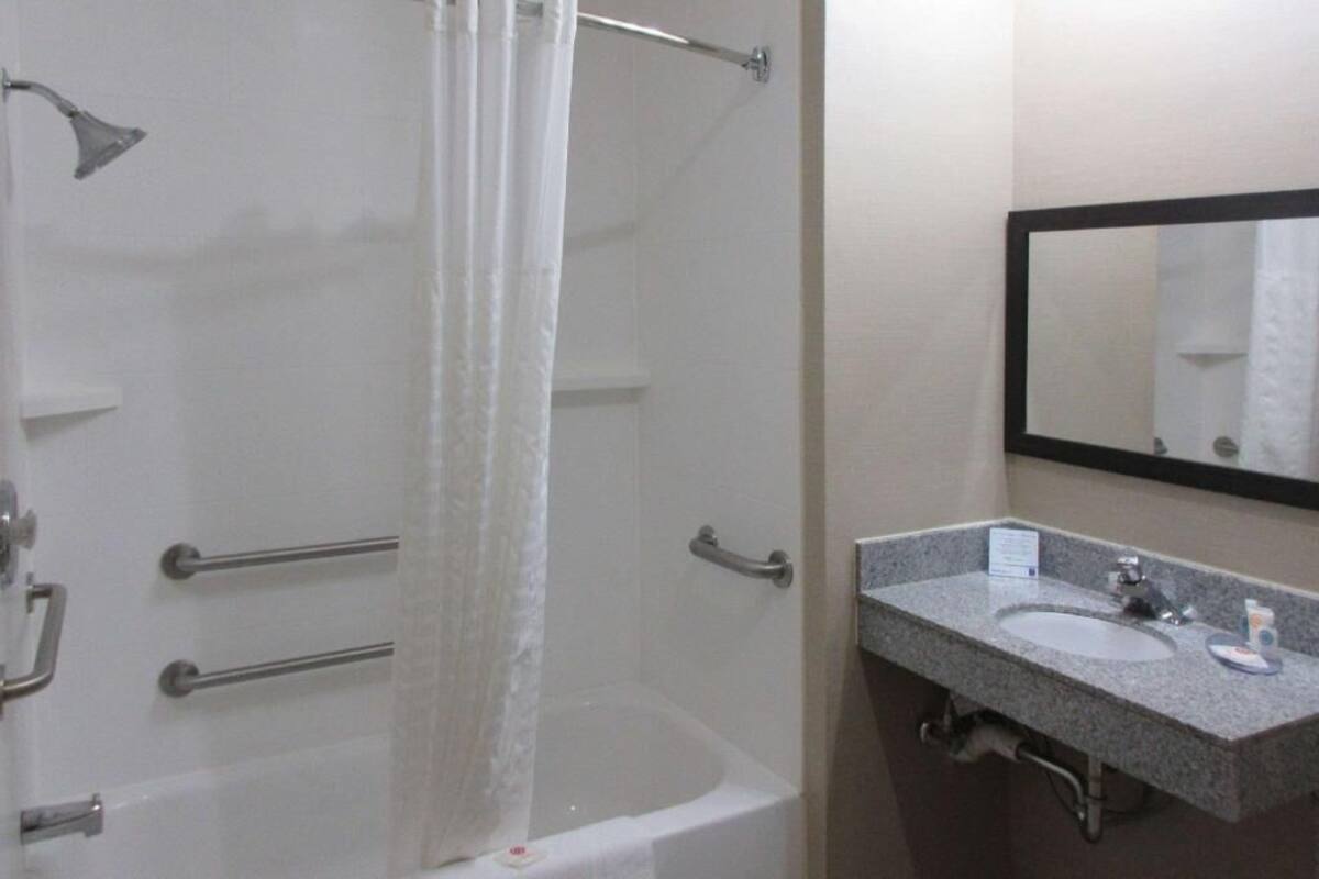 A bathroom with a bathtub, a shower curtain, a handrail, a sink, a mirror, and a towel rack.