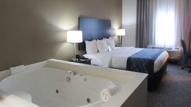 A hotel room with a large bathtub, a bed with a headboard, two bedside lamps, and a window with curtains. - Comfort Inn Port Huron (Port Huron)
