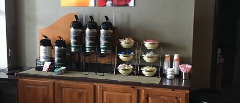 A breakfast bar with coffee dispensers, bowls of food, and a wall with pictures of food and a green sign.