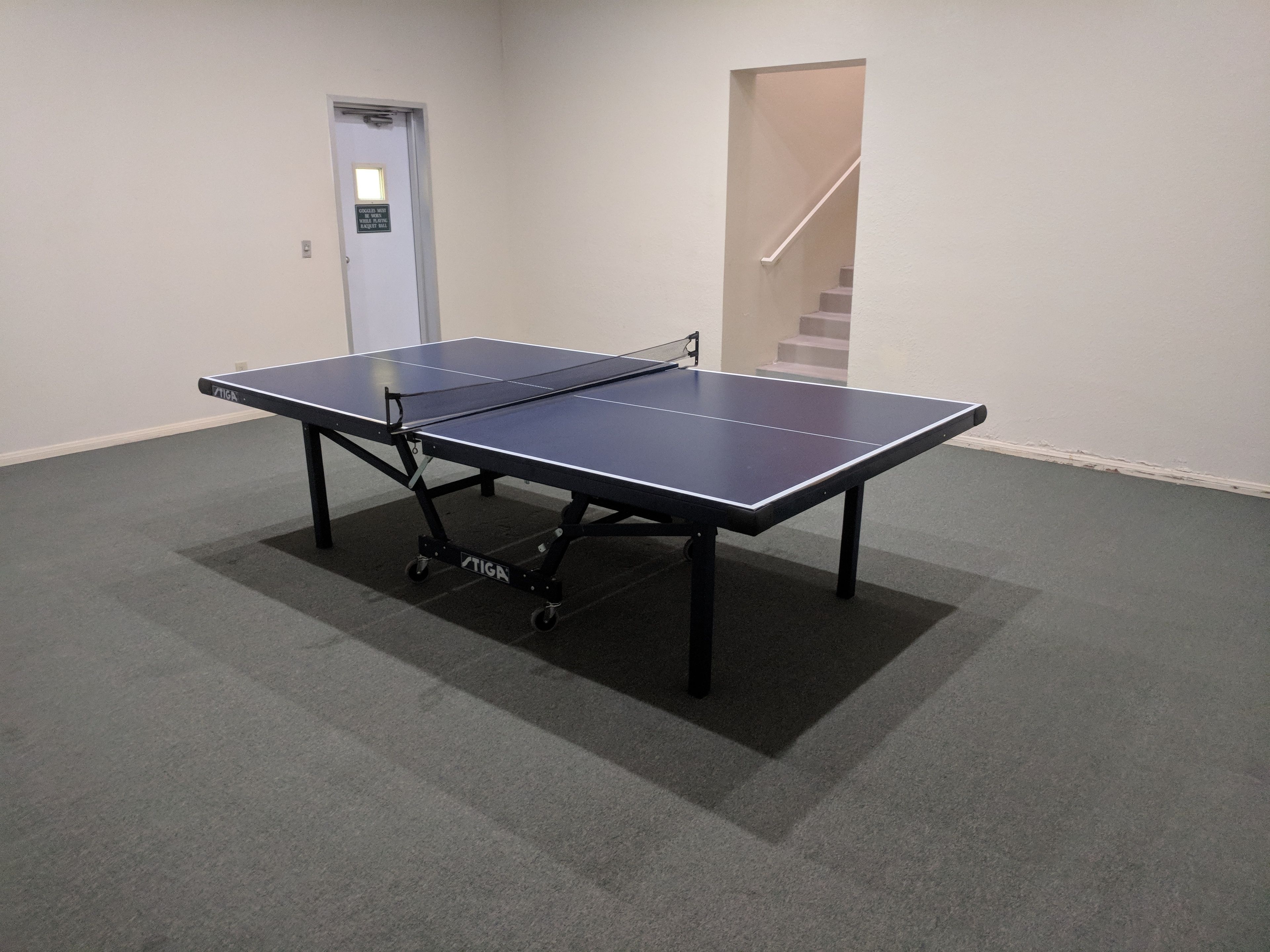 A ping pong table in a room with carpeted flooring and a door with a sign.