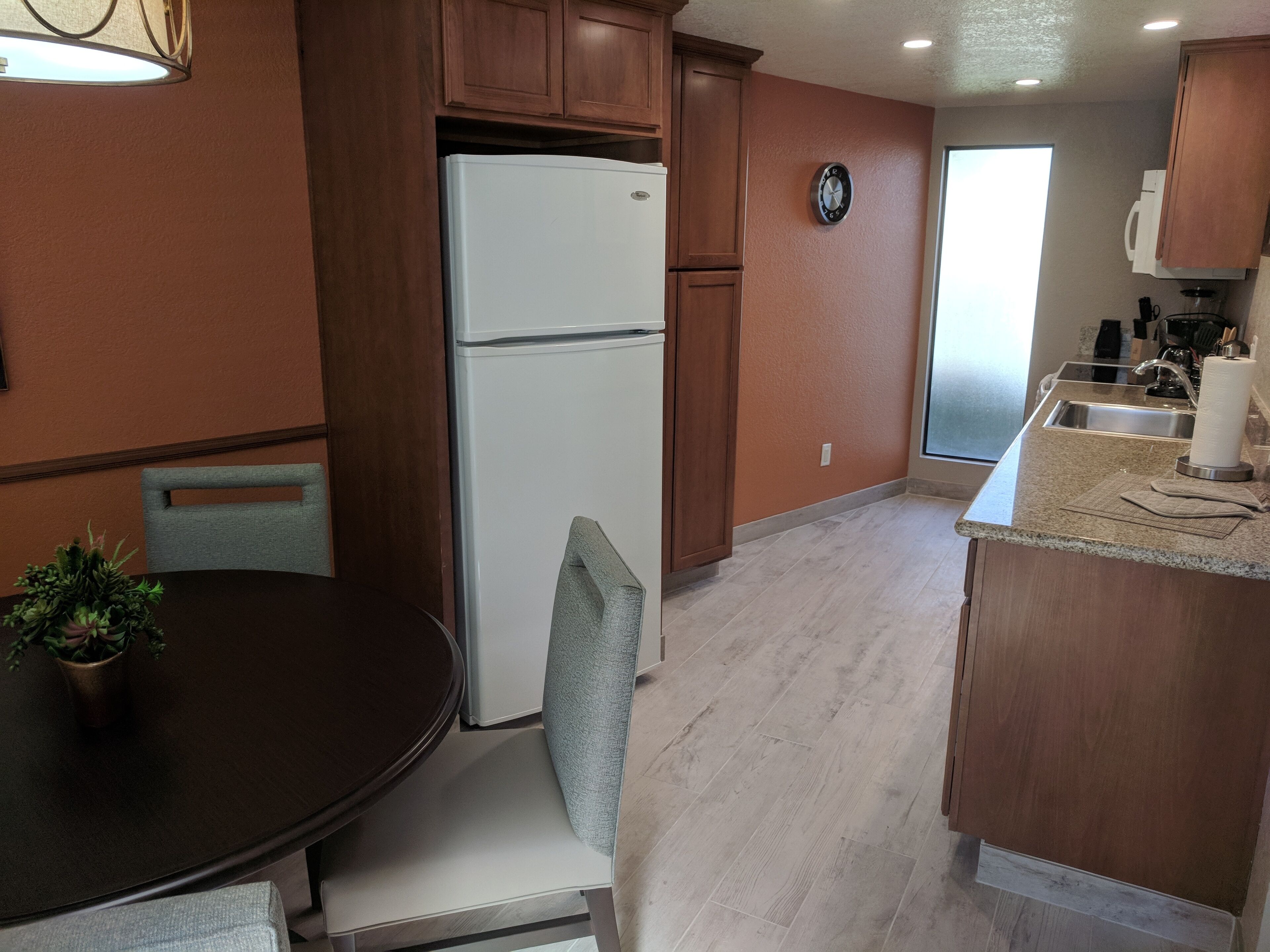 A compact kitchen with wooden cabinets, a white refrigerator, a clock on the wall, and a dining area with a table and chairs.