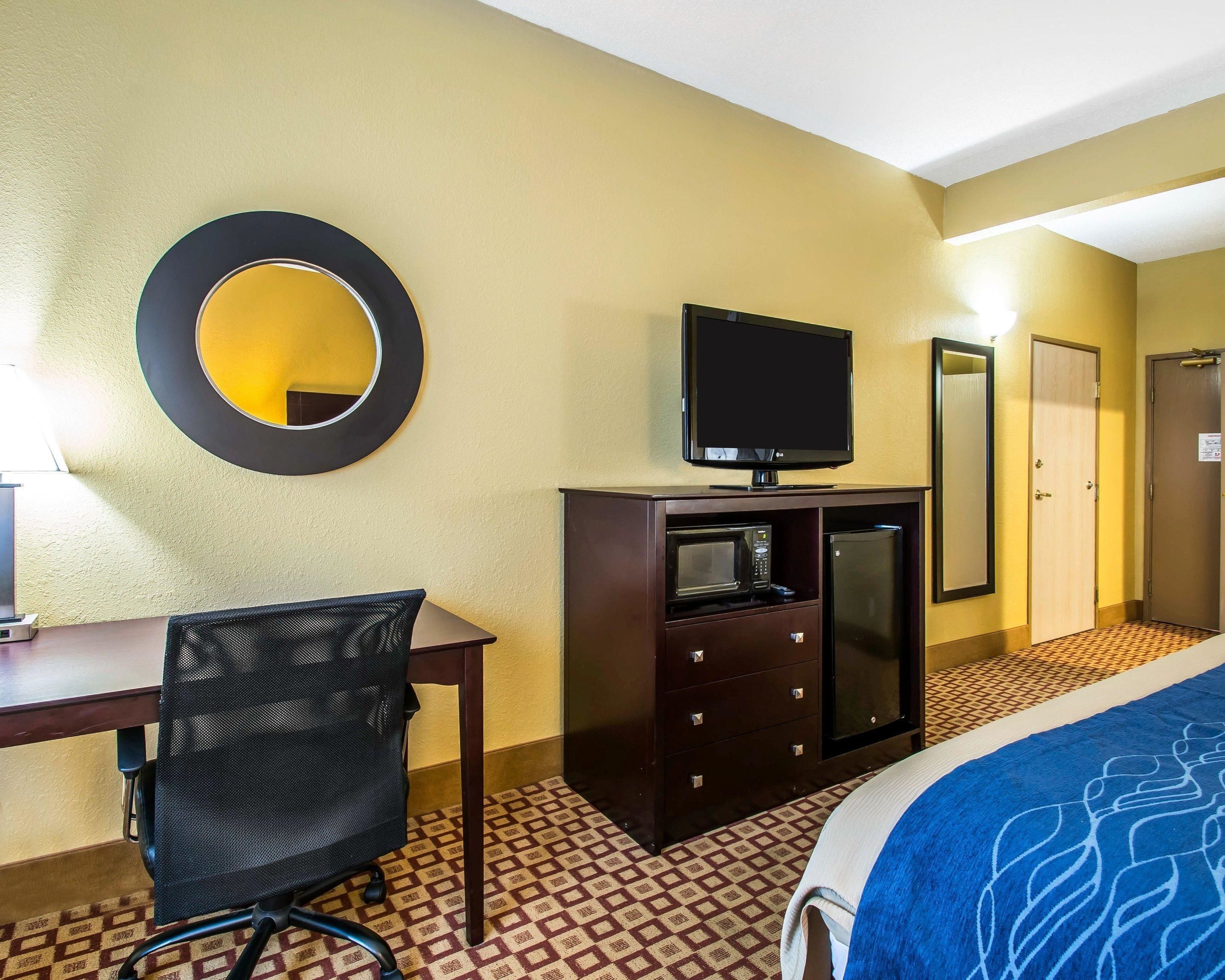 A hotel room with a bed, a desk with a chair, a television, a minibar, a mirror, and a door.
