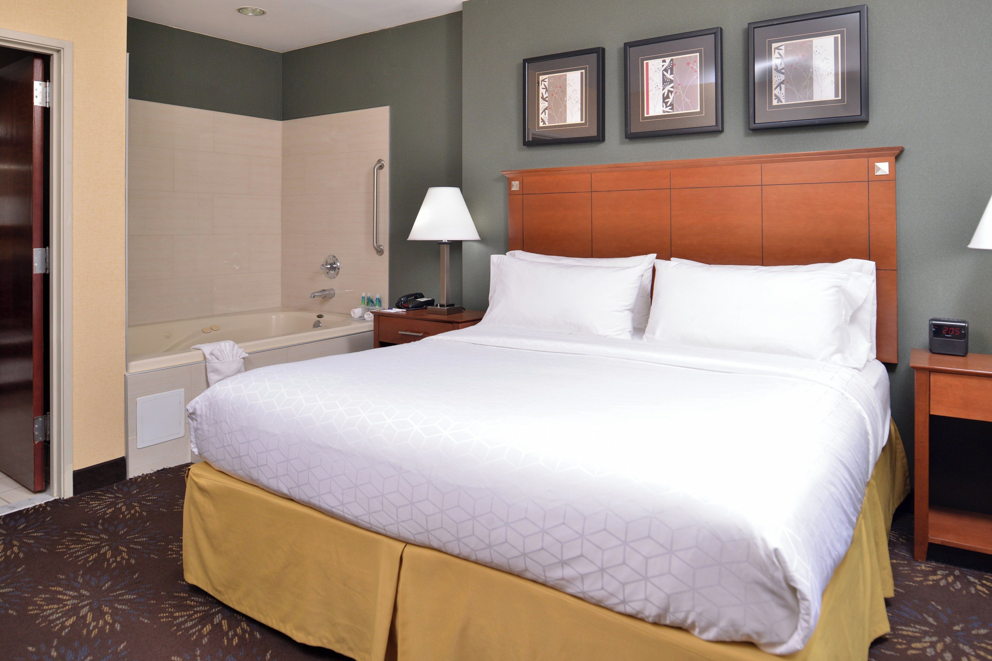 A hotel room with a large bed, a wooden headboard, a nightstand with a lamp, a bathroom with a bathtub, and a wall with framed artwork.