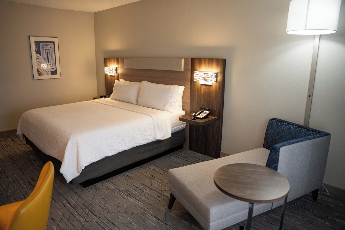 Quarto de hotel com uma cama grande, mesas de cabeceira, uma cadeira e uma pequena mesa.