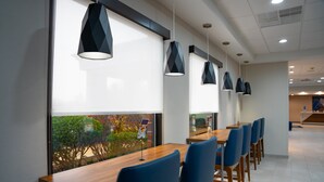 A modern dining area with a long wooden table and blue chairs, large windows with blinds, and pendant lights. - Holiday Inn Express Hotel & Suites West Chester by IHG (West Chester)
