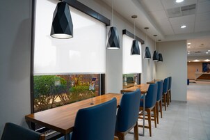Un comedor moderno con una larga mesa de madera y sillas azules, ventanas amplias con persianas e iluminación colgante.