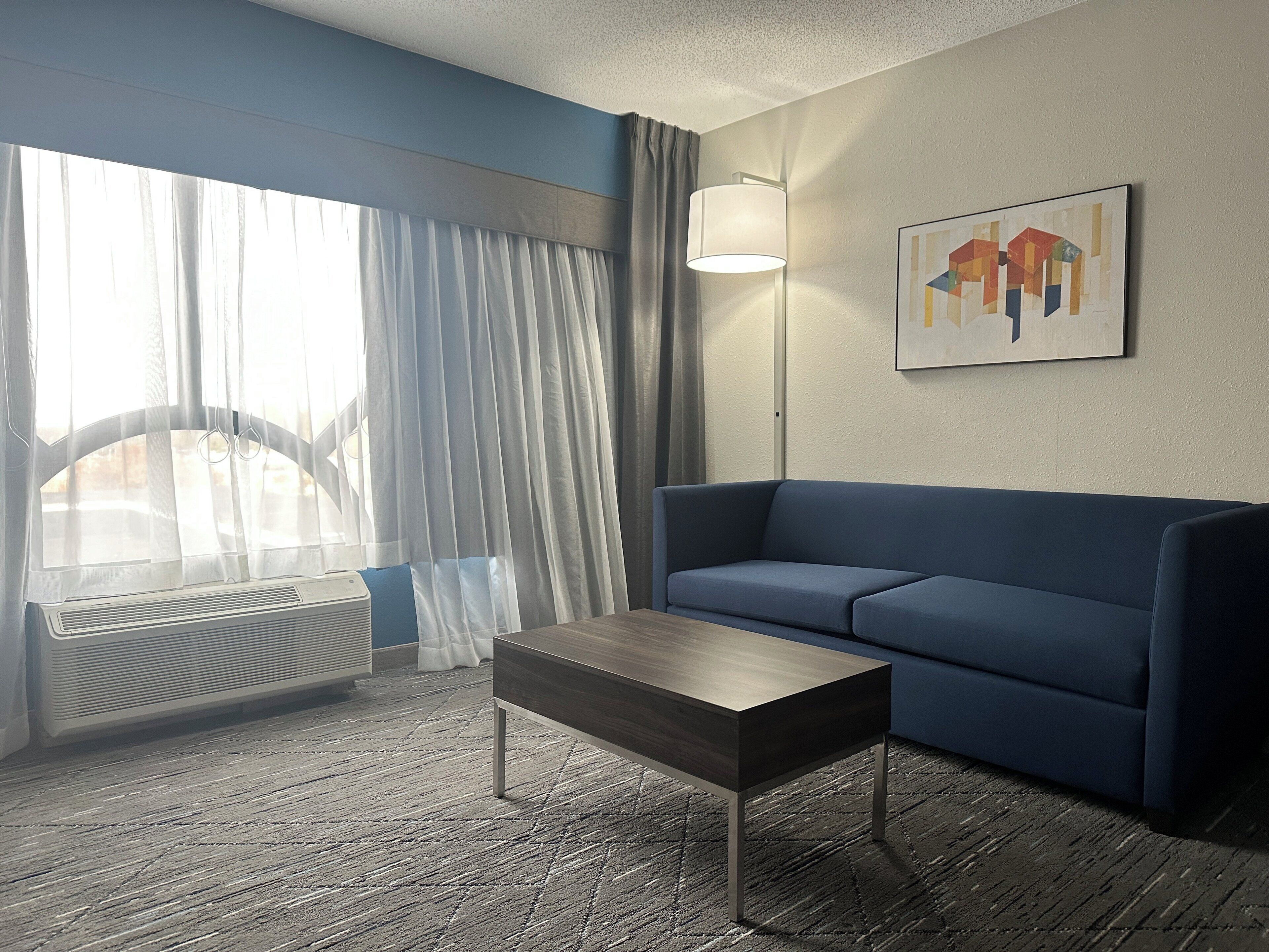 A hotel room with a blue sofa, a wooden coffee table, a floor lamp, a radiator, and a window with curtains.