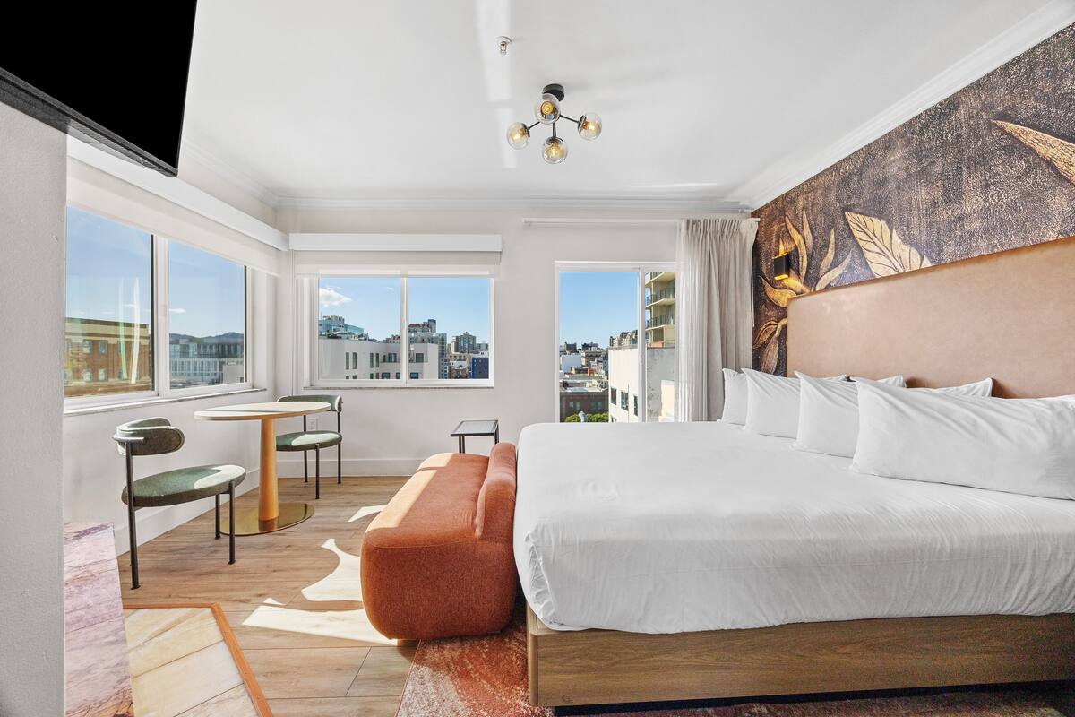 A modern hotel room with a large bed, a view of the city, a small table, and a sofa.