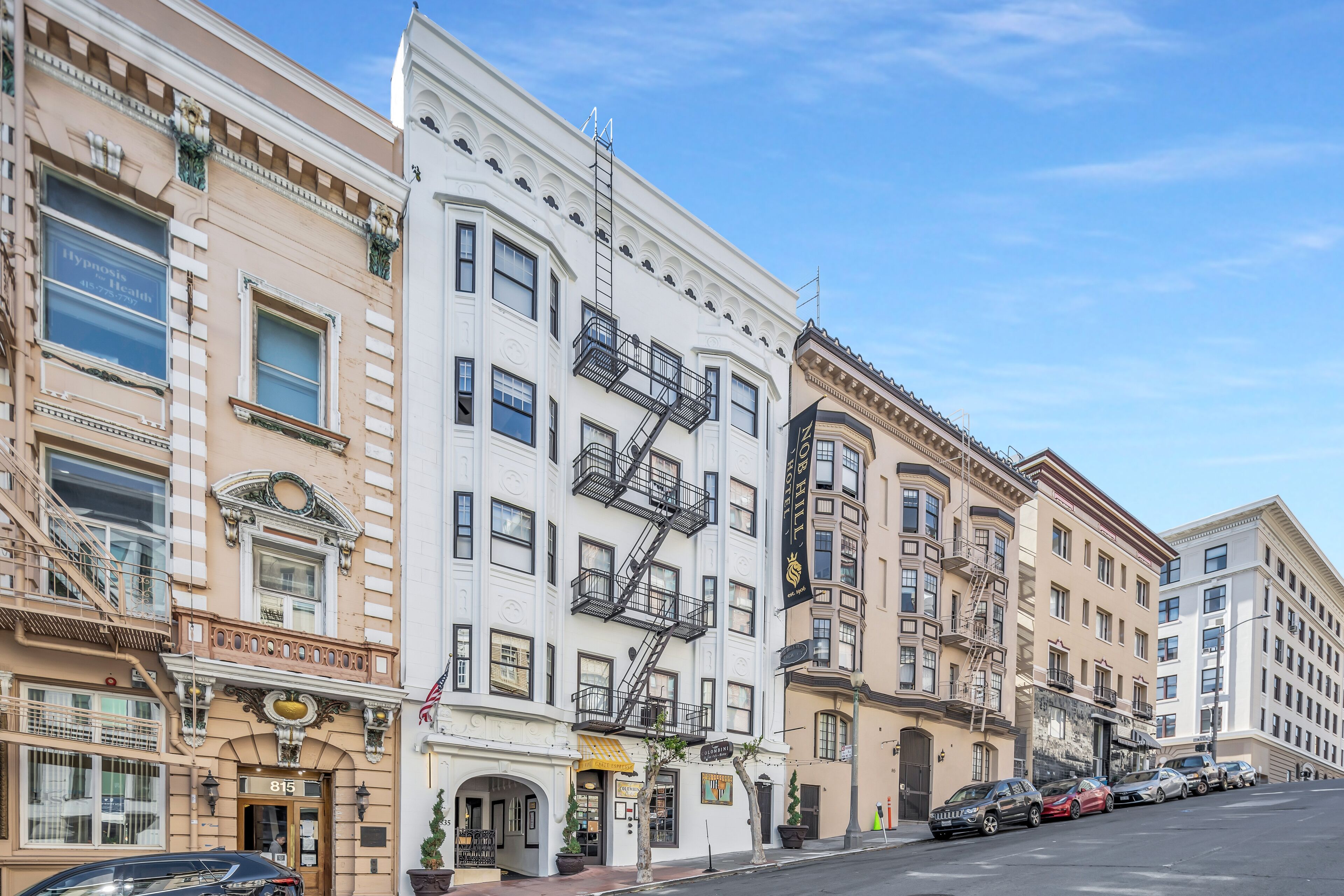 A row of multi-story buildings with fire escapes, a street, and parked cars.