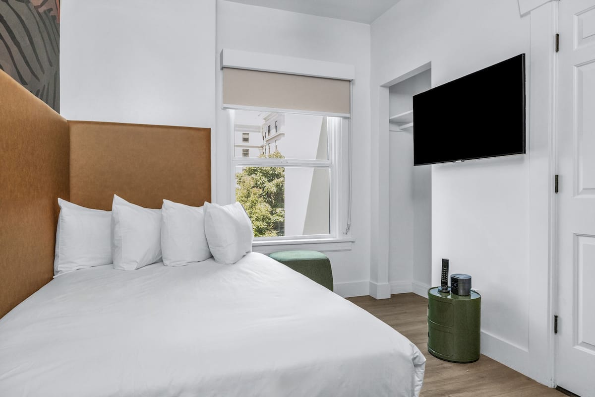A modern hotel room with a large bed, a television mounted on the wall, a window with a view of buildings, and a green side table.