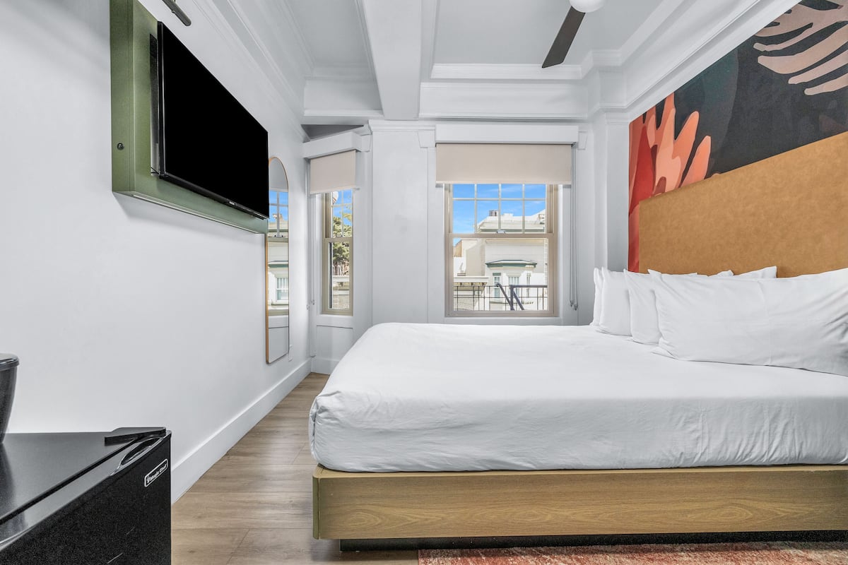 A modern hotel room with a large bed, a flat-screen TV mounted on the wall, and a view of the outside balcony.