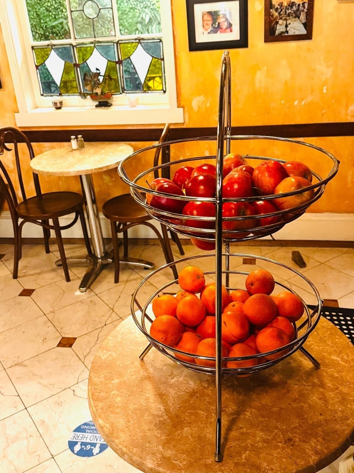 A dining area with a round table, two chairs, a metal fruit stand with apples and oranges, a window with curtains, and framed pictures on the wall.