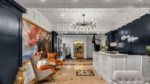 A lobby with a black reception desk, white walls, and a chandelier. There are two orange chairs and a white chair.