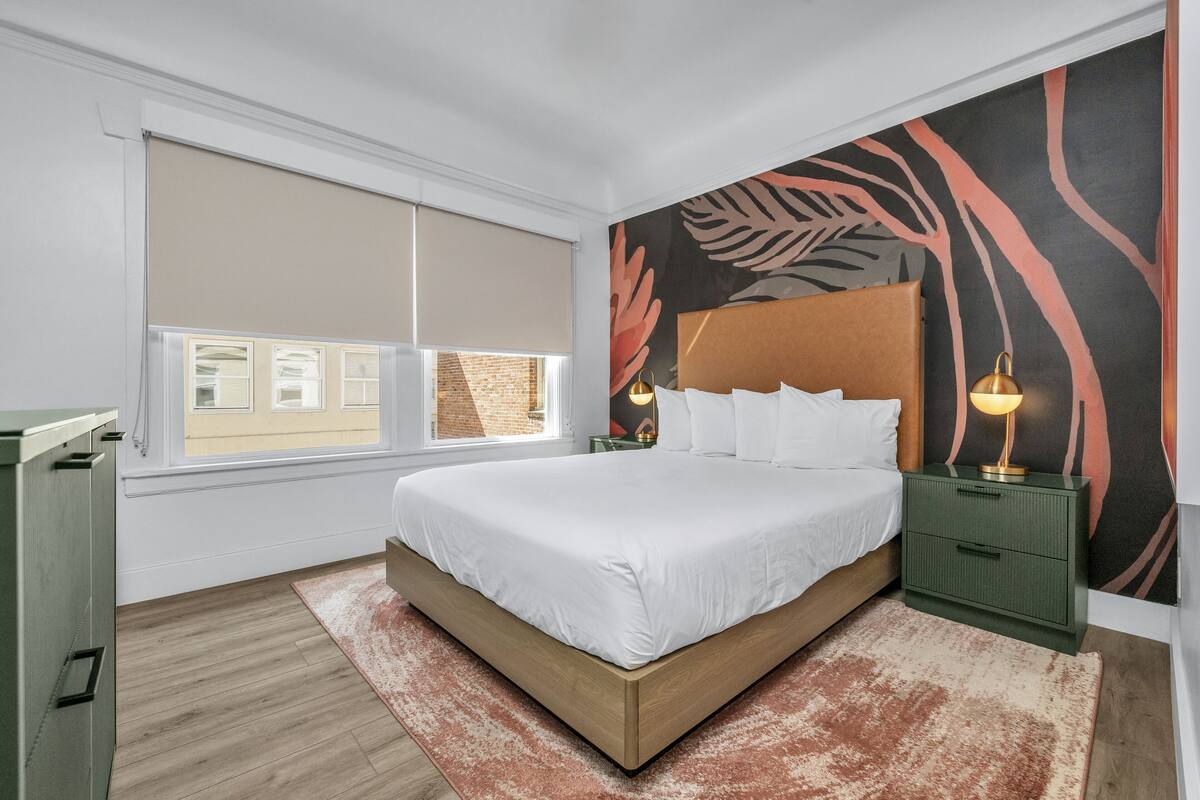 A bedroom with a bed, nightstands, a window with blinds, and a wall with a leaf pattern.