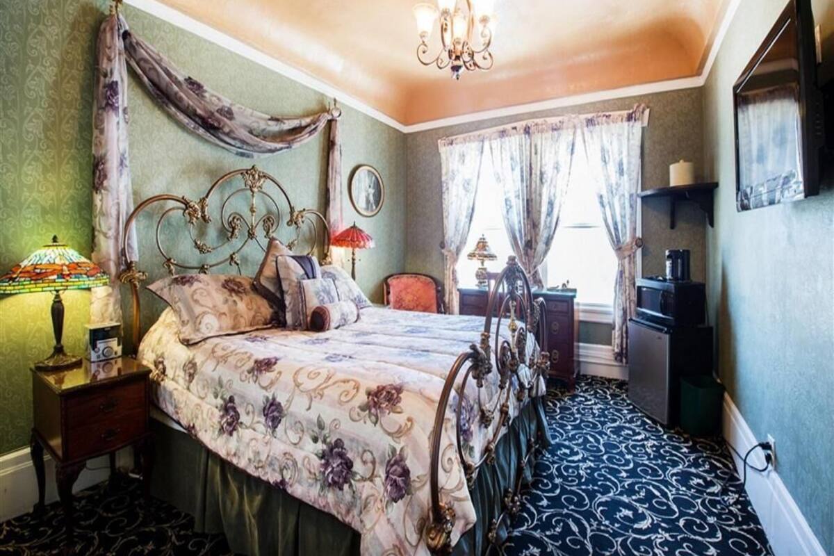 A bedroom with a four-poster bed, a chandelier, a desk with a lamp, and a television on the wall.
