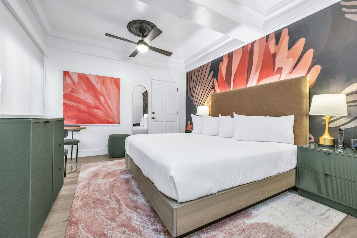 A modern hotel room with a large bed, a ceiling fan, a green nightstand, and a vibrant abstract painting on the wall.