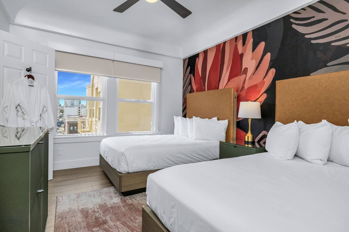 A hotel room with two beds, a large window with a city view, a green dresser, and a wall with a large abstract painting.