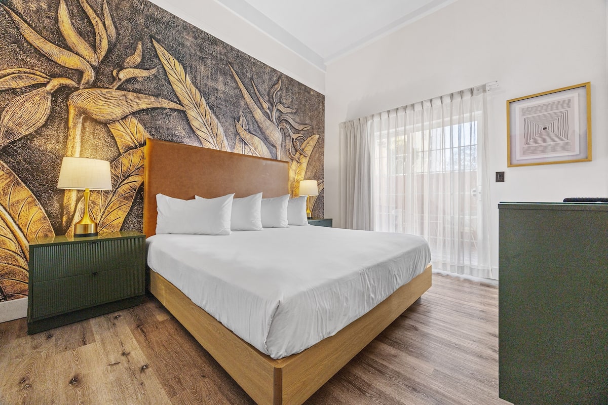 A hotel room with a large bed, two bedside tables with lamps, a wall mural, and a window with curtains.