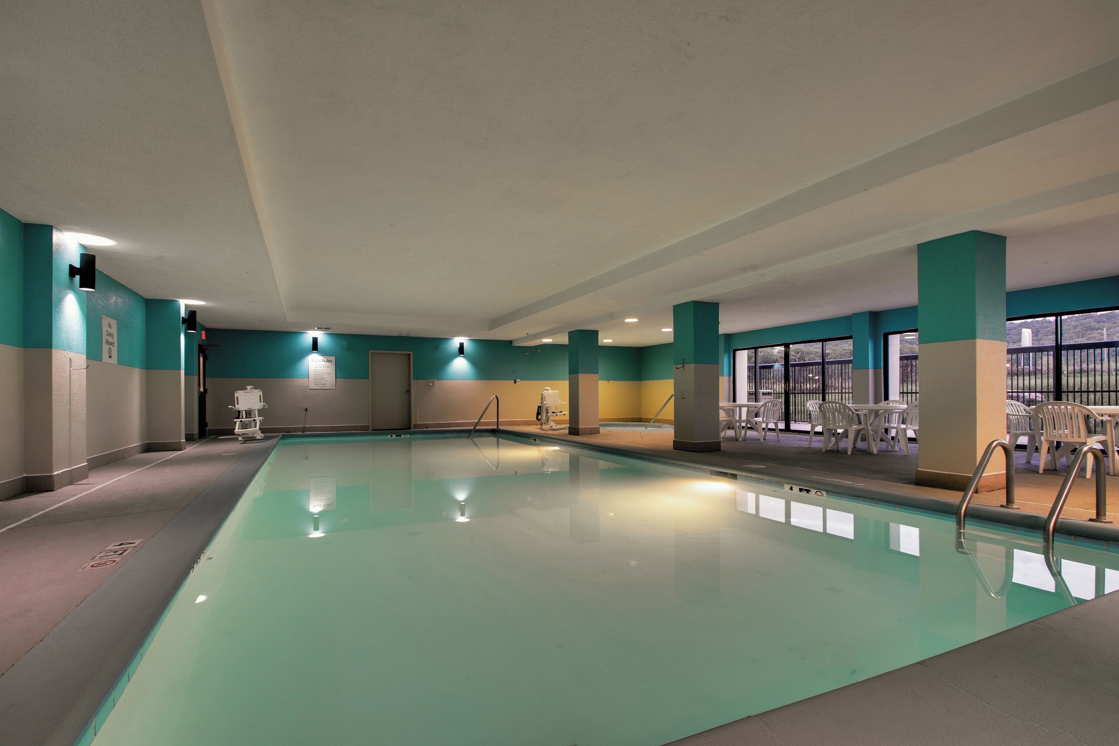 Una piscina interior con agua cristalina, rodeada de paredes en tonos verde azulado y beige, y un área de descanso con sillas blancas.