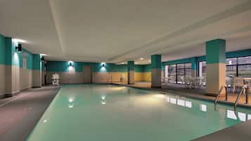 Una piscina interior con agua cristalina, rodeada de paredes en tonos verde azulado y beige, y un área de descanso con sillas blancas.