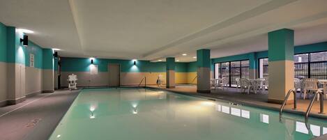Una piscina interior con agua cristalina, rodeada de paredes en tonos verde azulado y beige, y un área de descanso con sillas blancas.