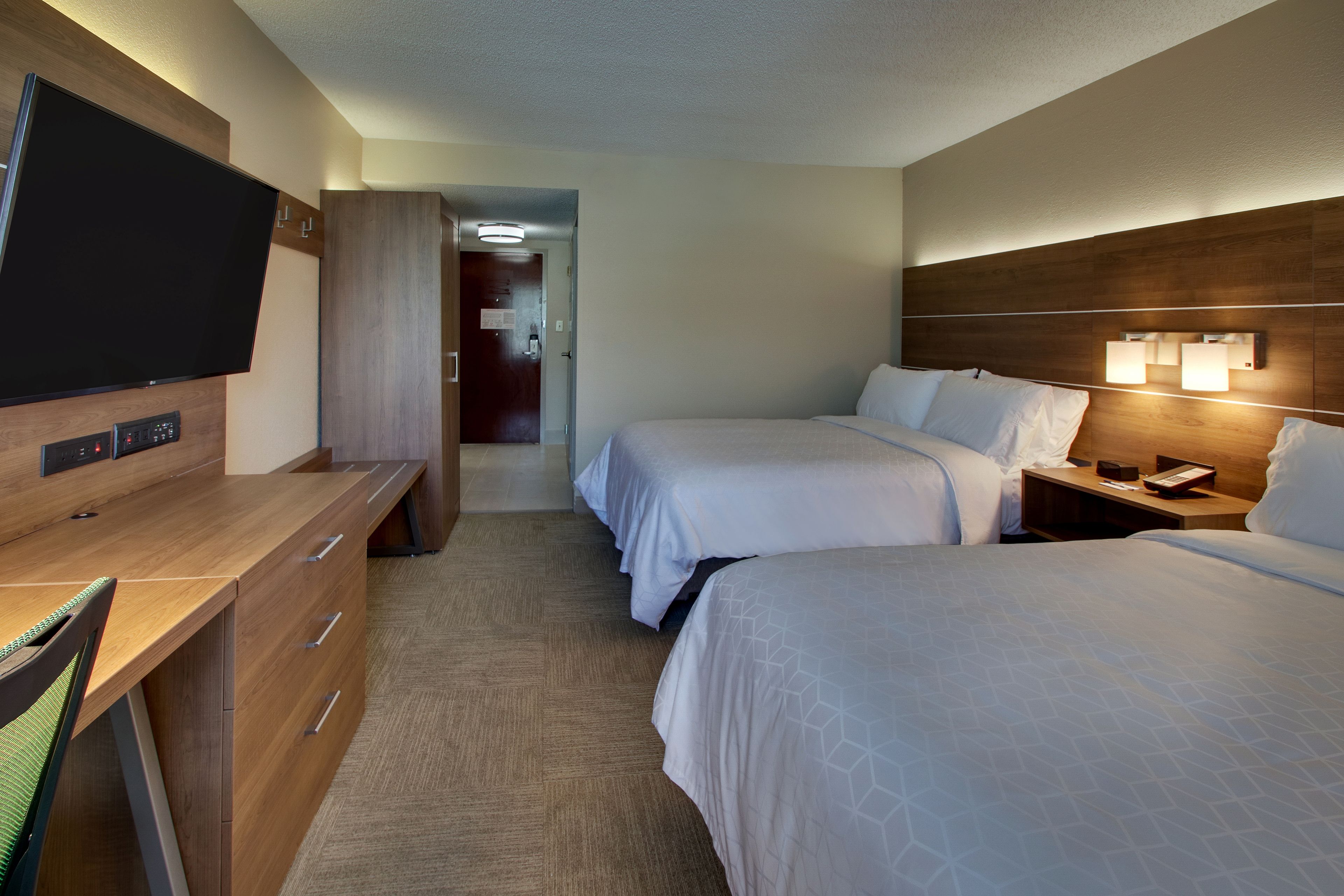 A hotel room with two beds, a desk with a flat-screen TV, a dresser, and a nightstand with a lamp.