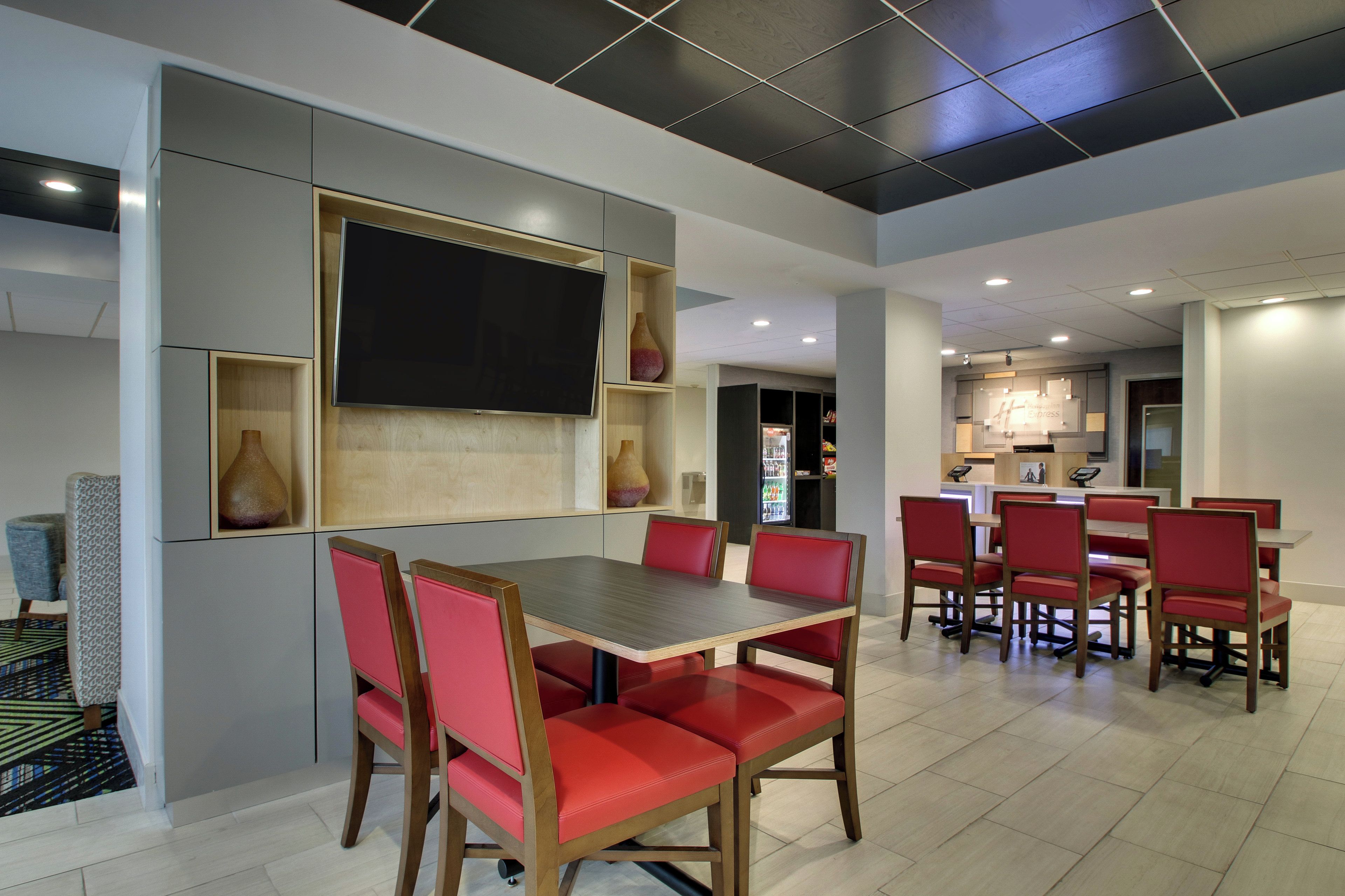 A modern dining area with a large table and red upholstered chairs, a flat-screen TV mounted on the wall, and a kitchen in the background.