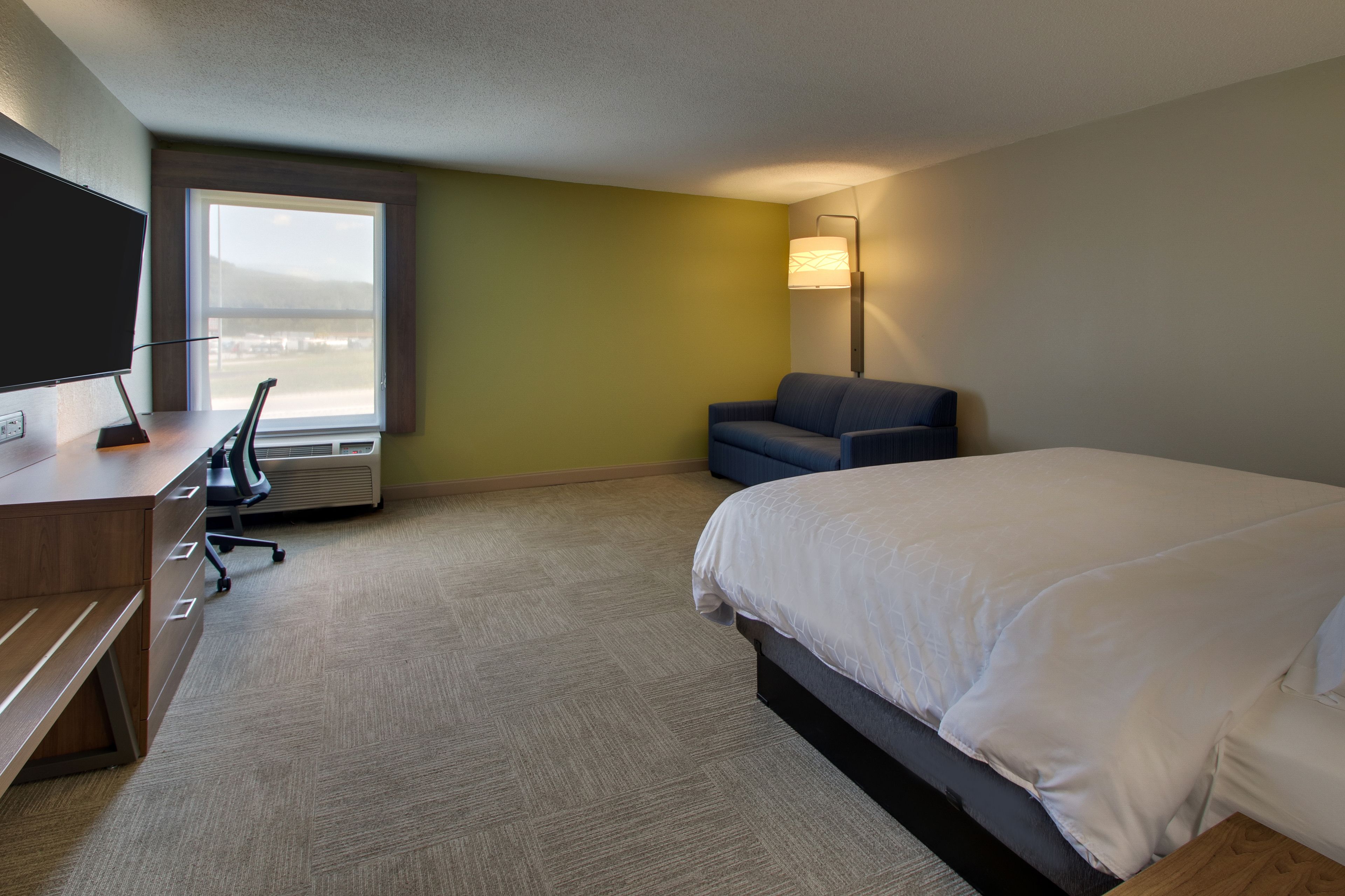 A hotel room with a large bed, a desk with a chair, a TV, a lamp, and a window with a view.