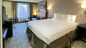 Une chambre d’hôtel avec un grand lit, un bureau, une chaise, une lampe de bureau, une table de chevet et une fenêtre avec des rideaux.